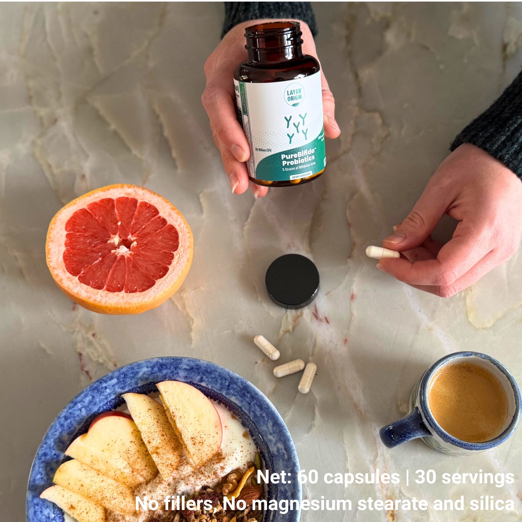 Bifidobacteria Person holding a bottle of supplements with a half-sliced grapefruit, apple slices, and a cup of coffee on a marble surface.
